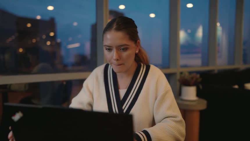A young woman works diligently on her laptop in a modern office at dusk, taking a break to sip coffee before smiling at her screen