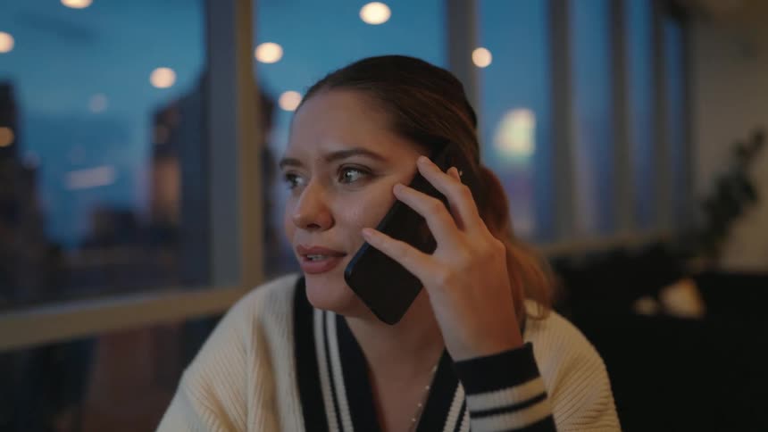 A happy young woman sitting by a window with a city view at dusk answers her phone and has an engaging conversation