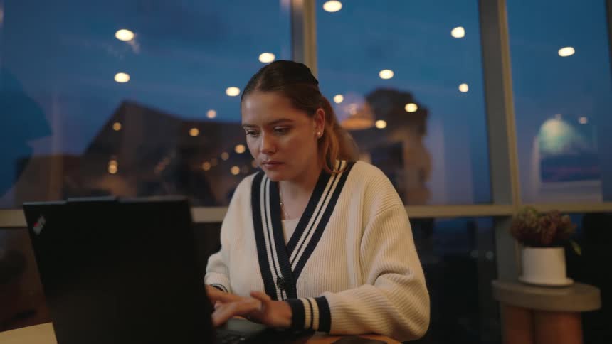 A young woman sips coffee and works on her laptop at a desk in front of a window in the evening