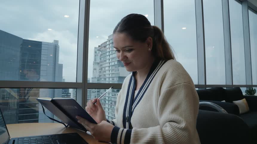 A smiling young professional woman works on her laptop and takes notes in her journal in a modern office with a city skyline view