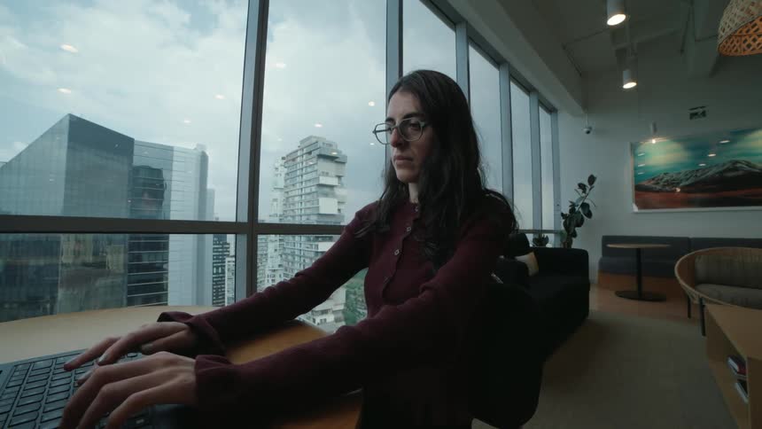 A young professional woman with glasses types on her laptop in a modern high-rise office with a city view, as the camera slowly pulls back