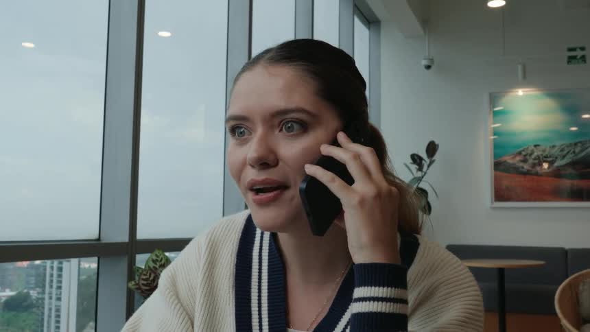 A happy young woman's expression shifts to joyful surprise as she takes an exciting phone call in a modern cafe overlooking the city