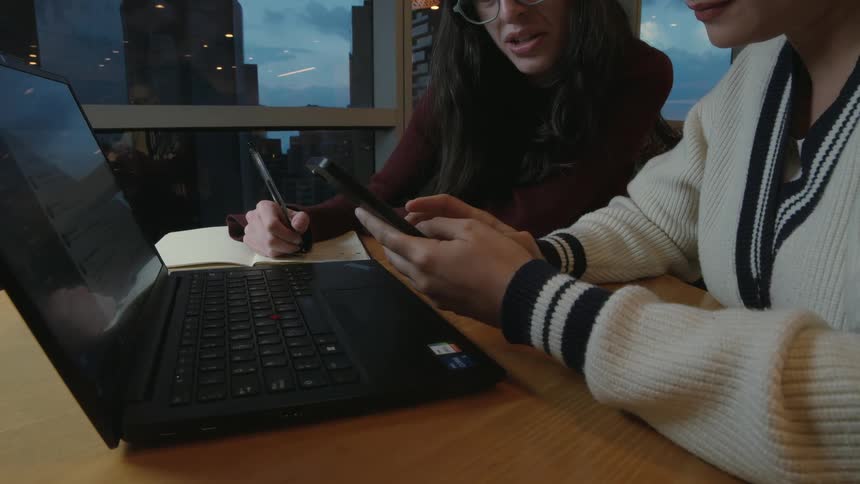 Two young women happily collaborate on a project in a city cafe, sharing ideas from a smartphone while one takes notes