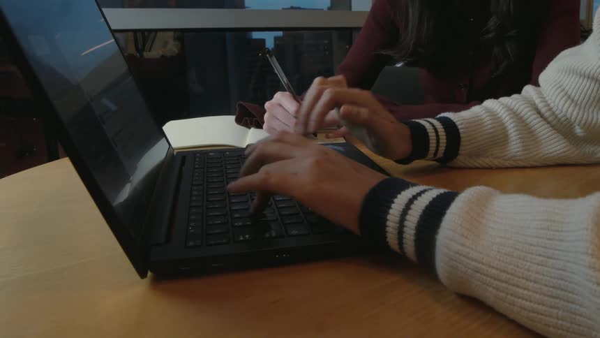 A close-up of two people working together at a table, one typing on a laptop while the other writes notes in a notebook