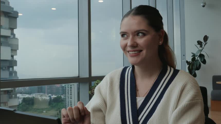 Close-up of a young professional woman smiling as she works on her laptop by a large window overlooking the city