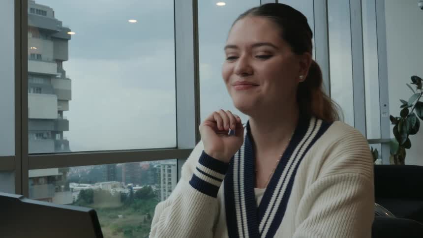 A smiling young woman waves during a video call on her laptop in a modern office with a city view