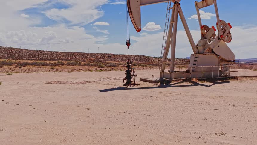 An oil pumpjack steadily works to extract crude oil from a well in the vast, arid desert under a bright blue sky