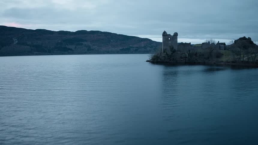 An aerial view slowly approaches the ancient ruins of a stone castle perched on the shore of a vast lake at dusk