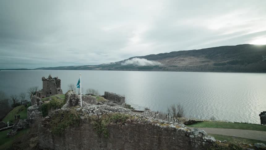 A cinematic aerial shot rises over the ancient stone walls of Urquhart Castle, revealing the famous Loch Ness in Scotland under a cloudy sky