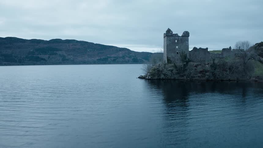 Flying low over the dark waters of Loch Ness towards the ancient ruins of Urquhart Castle on a cloudy day in Scotland