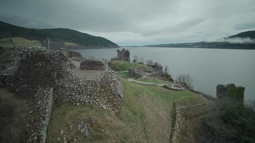 A slow reveal of the ancient stone ruins of Urquhart Castle on the banks of Loch Ness in Scotland under a cloudy sky