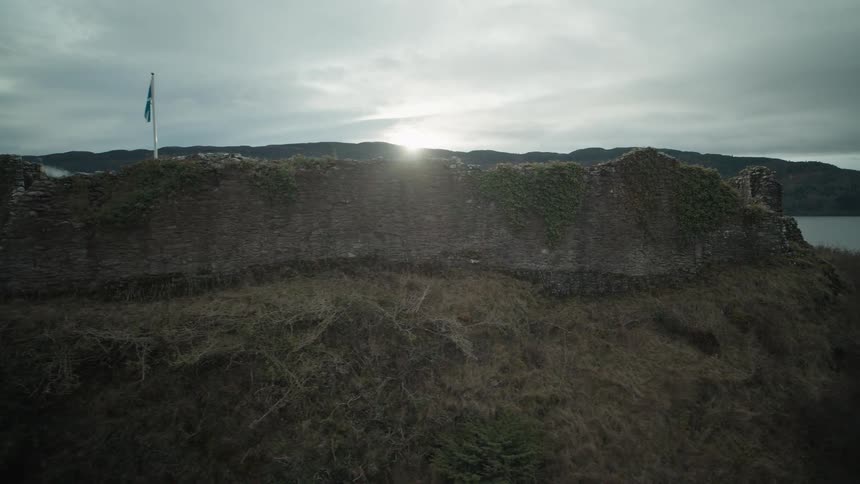 A slow drone shot reveals the historic ruins of Urquhart Castle on the banks of Loch Ness, with the Scottish flag flying against a moody, overcast sunset