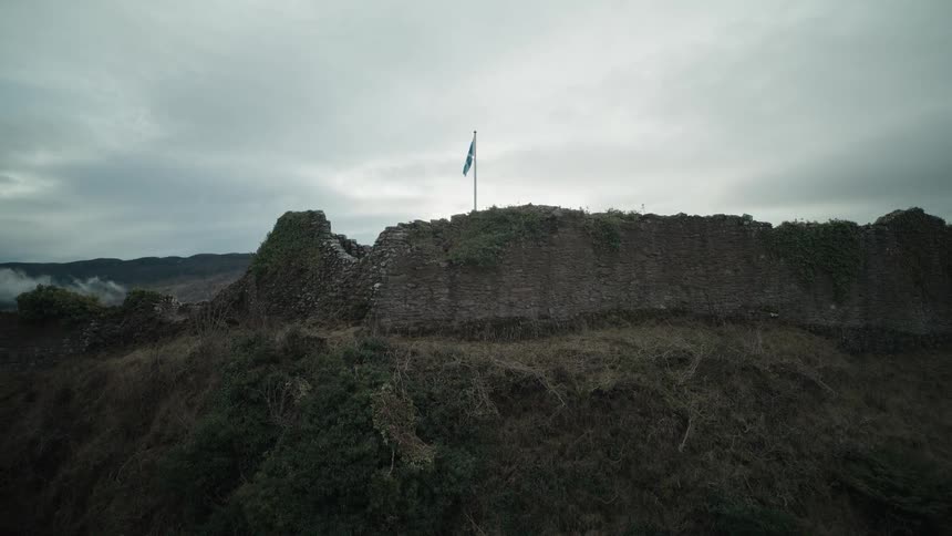 Dramatic aerial reveal of the Scottish flag on the ruins of Urquhart Castle overlooking Loch Ness as the sun breaks through the clouds