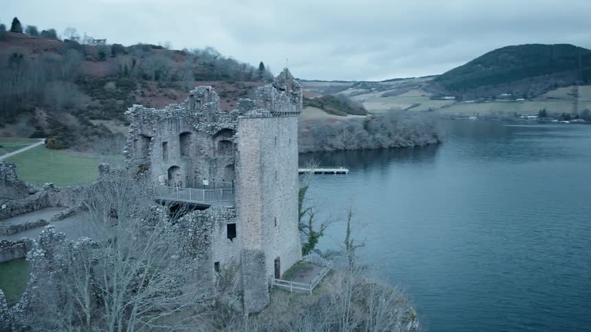 A sweeping aerial view reveals the ancient and historic Urquhart Castle ruins overlooking the vast expanse of Loch Ness in the Scottish Highlands