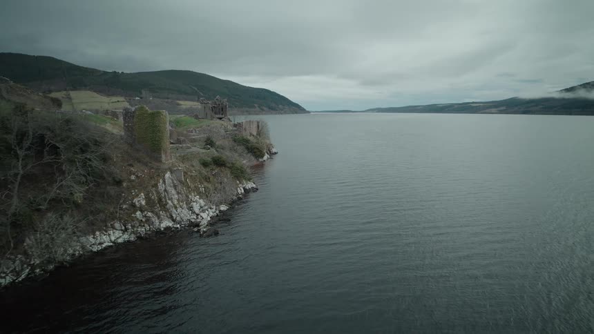 A slow aerial approach reveals the historic Urquhart Castle ruins on the shores of Loch Ness in Scotland on a moody, overcast day