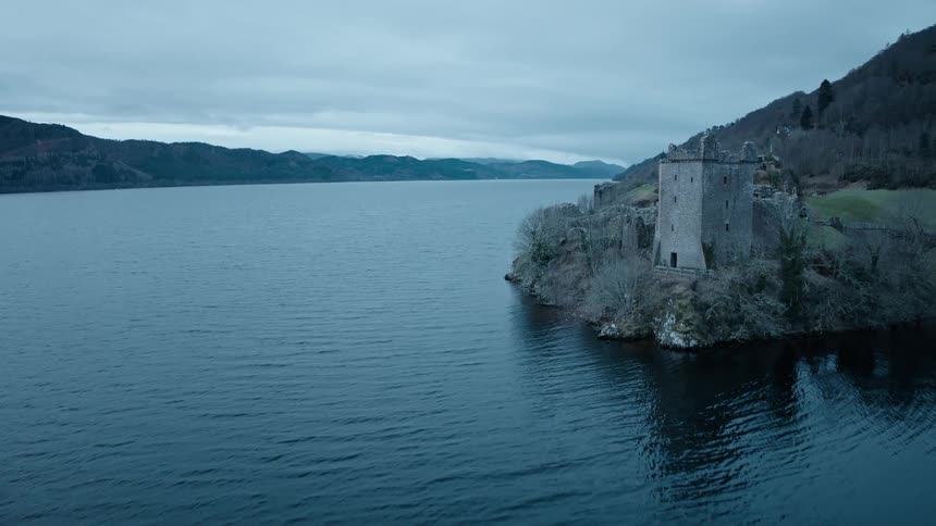 A slow aerial flight over the dark waters of Loch Ness reveals the historic ruins of Urquhart Castle on a moody, overcast day in the Scottish Highlands