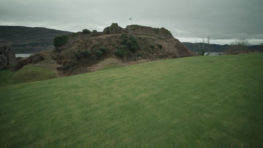 A sweeping aerial view travels over a green hill towards the ancient stone ruins of Urquhart Castle, where a Scottish flag flies over the waters of Loch Ness