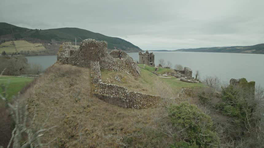 An aerial view slowly pushes in on the historic Urquhart Castle ruins overlooking Loch Ness, revealing a bagpiper in a traditional kilt