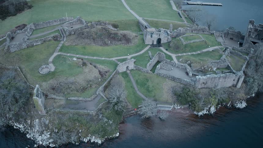 A slow aerial pan showcases the ancient and sprawling ruins of Urquhart Castle overlooking the dark waters of Loch Ness in the Scottish Highlands