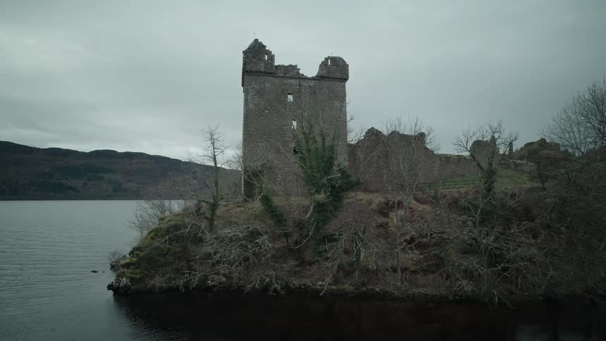 A cinematic aerial shot slowly pans left around the historic ruins of Urquhart Castle on the banks of Loch Ness in Scotland