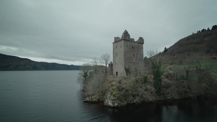 An aerial view pulls back from the ancient ruins of Urquhart Castle on the shores of Loch Ness on a moody, overcast day in the Scottish Highlands