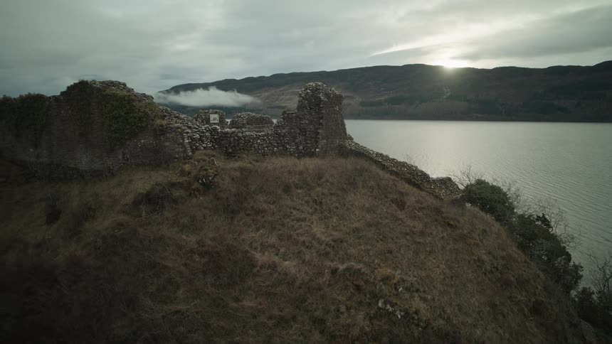A dramatic aerial reveal of the historic Urquhart Castle ruins overlooking Loch Ness in the Scottish Highlands at sunset