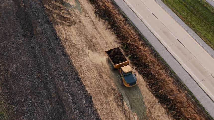Aerial top-down shot of an excavator filling a dump truck with soil at a highway construction site before the truck drives off