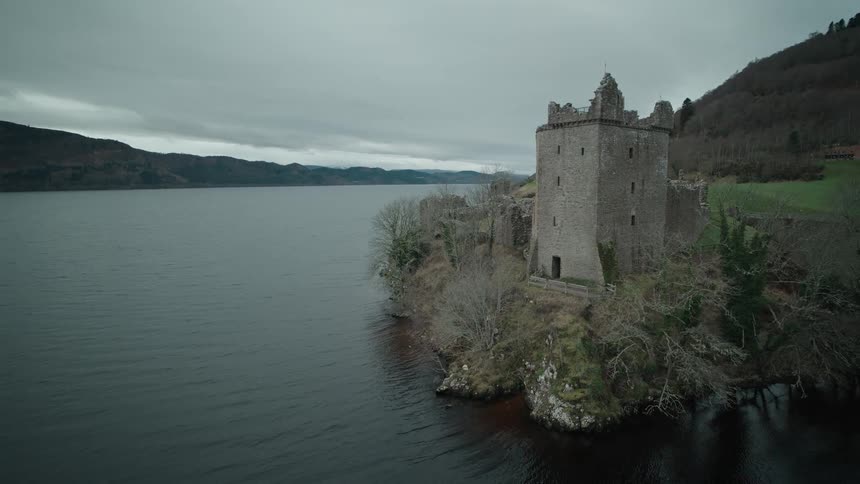 An aerial pullback reveals the ancient ruins of Urquhart Castle overlooking the dark waters of Loch Ness in Scotland