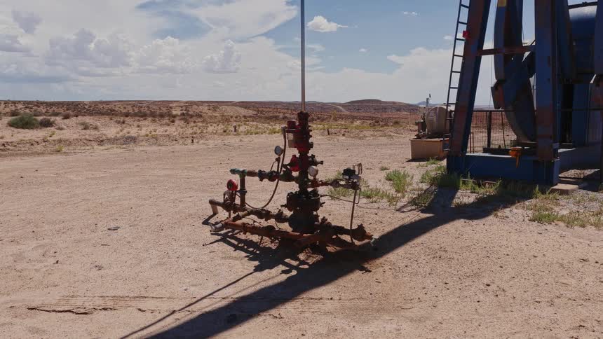 An oil pumpjack operates tirelessly, extracting crude oil from a wellhead in the middle of a vast, arid desert under a bright, partly cloudy sky