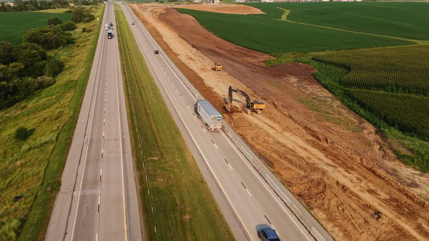 Aerial footage captures cars and semi-trucks on a rural highway alongside a major road construction project with heavy machinery