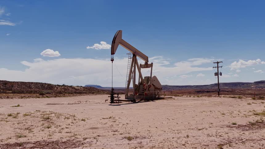 An oil pumpjack, or nodding donkey, continuously pumps crude oil in a vast, arid desert landscape under a bright blue sky