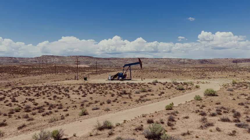 An aerial view slowly advances on a working oil pumpjack in a desolate desert environment with distant mesas