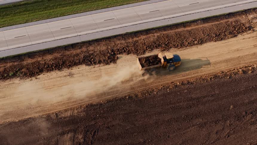 High-angle aerial tracking shot of a large articulated dump truck driving along a dirt road at a construction site, kicking up dust next to a busy highway