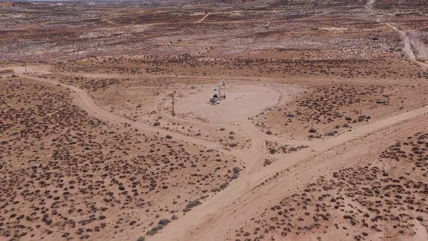 An aerial view flies towards a solitary oil pumpjack operating in a vast and arid desert landscape