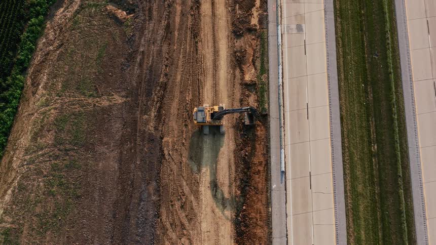 An aerial top-down view shows an excavator at a construction site working alongside a busy highway with passing cars and trucks