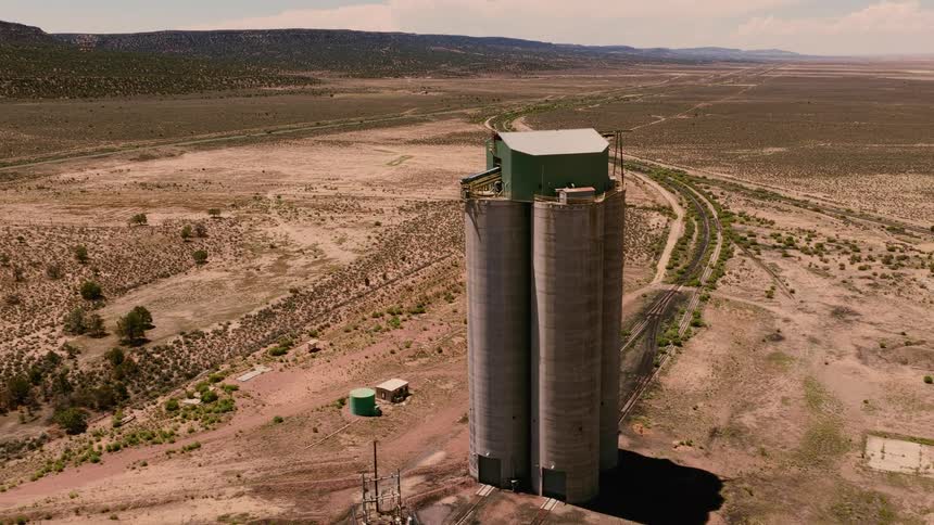 A slow aerial pullback reveals large industrial grain silos standing alone next to railroad tracks in a vast desert landscape