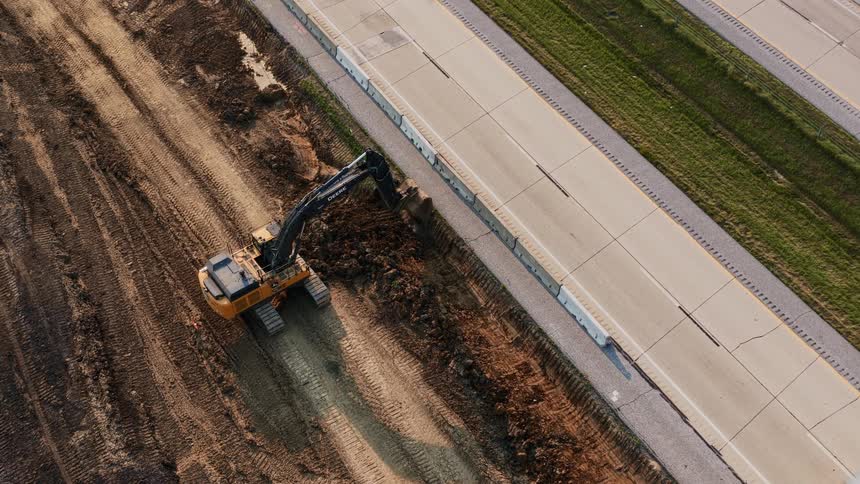 An aerial top-down view of a large excavator digging a trench on a construction site next to a busy highway with steady traffic flow