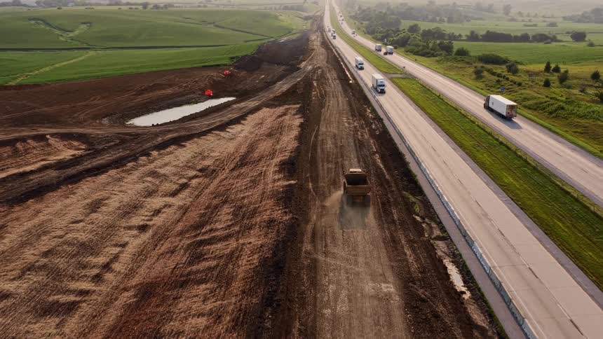 Aerial view of a large dump truck driving on a construction site alongside a busy highway through rural farmland