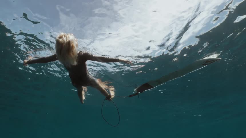 A serene underwater cinemagraph captures a blonde surfer in a wetsuit floating peacefully on the surface of the ocean
