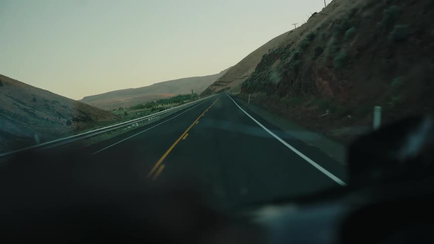 A point-of-view shot from inside a car traveling along a scenic highway winding through rolling hills at dawn