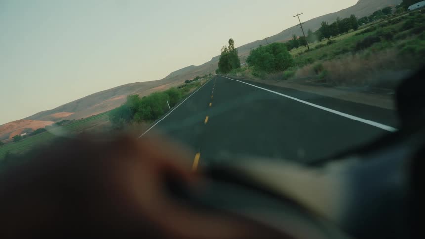 A serene first-person view from inside a car driving along a rural road surrounded by hills and fields at dusk
