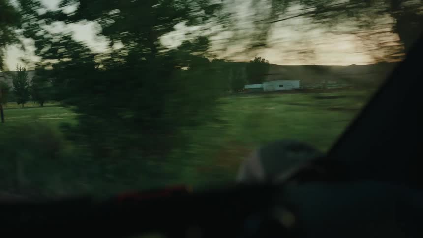 Driving through the serene countryside at dusk, with a view of green fields and a distant farm from the car window