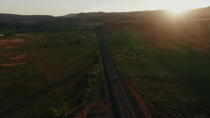 A lone car drives down a long, straight highway through a vast, green valley as the sun sets over the hills