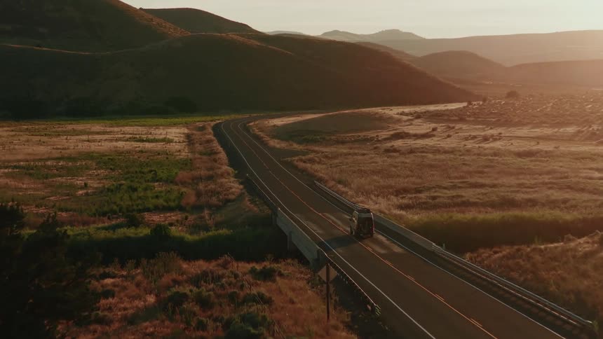Aerial view of a camper van driving across a bridge on a winding road through a golden, hilly landscape at sunset