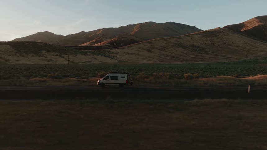 Aerial shot of a camper van driving down a desert highway surrounded by mountains at golden hour