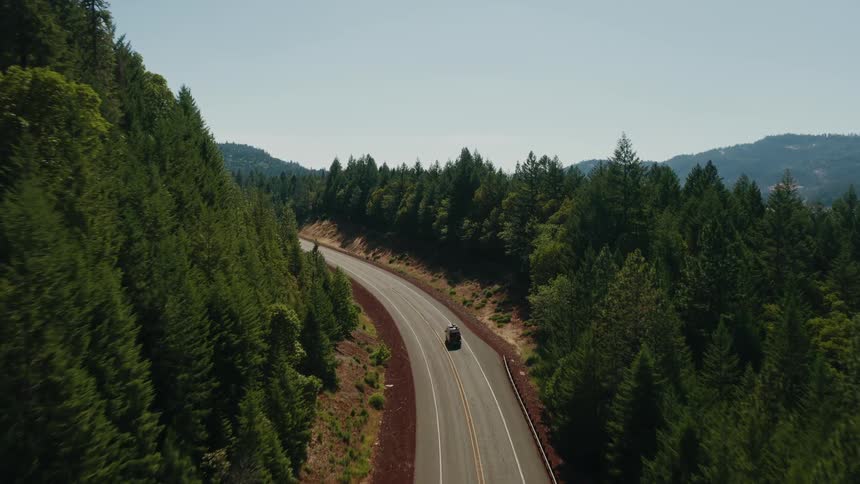 An aerial view follows a lone car driving on a scenic, winding road through a dense evergreen forest on a sunny day