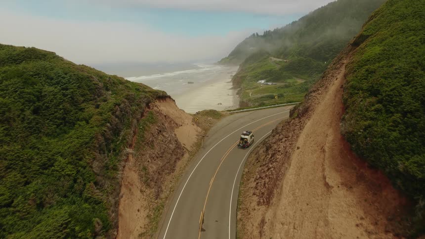 Aerial view of a camper van driving along a winding coastal highway carved into lush green cliffs with the ocean below