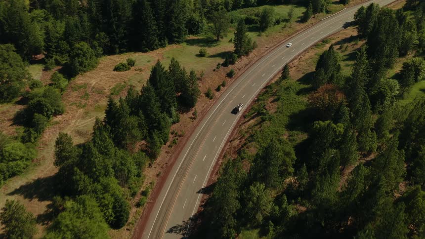 An aerial view shows a silver convertible driving along a winding country road surrounded by a lush green forest on a sunny day