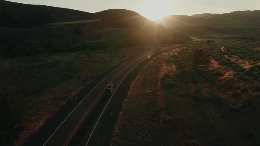 An aerial shot captures a vehicle driving along a winding highway through a vast, hilly landscape at golden hour