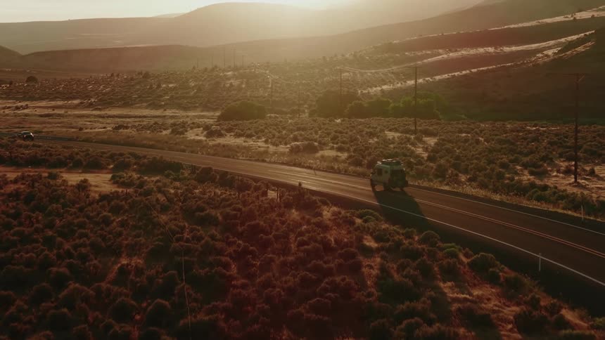 An aerial view follows a camper van driving on a remote desert road during a beautiful golden hour sunset